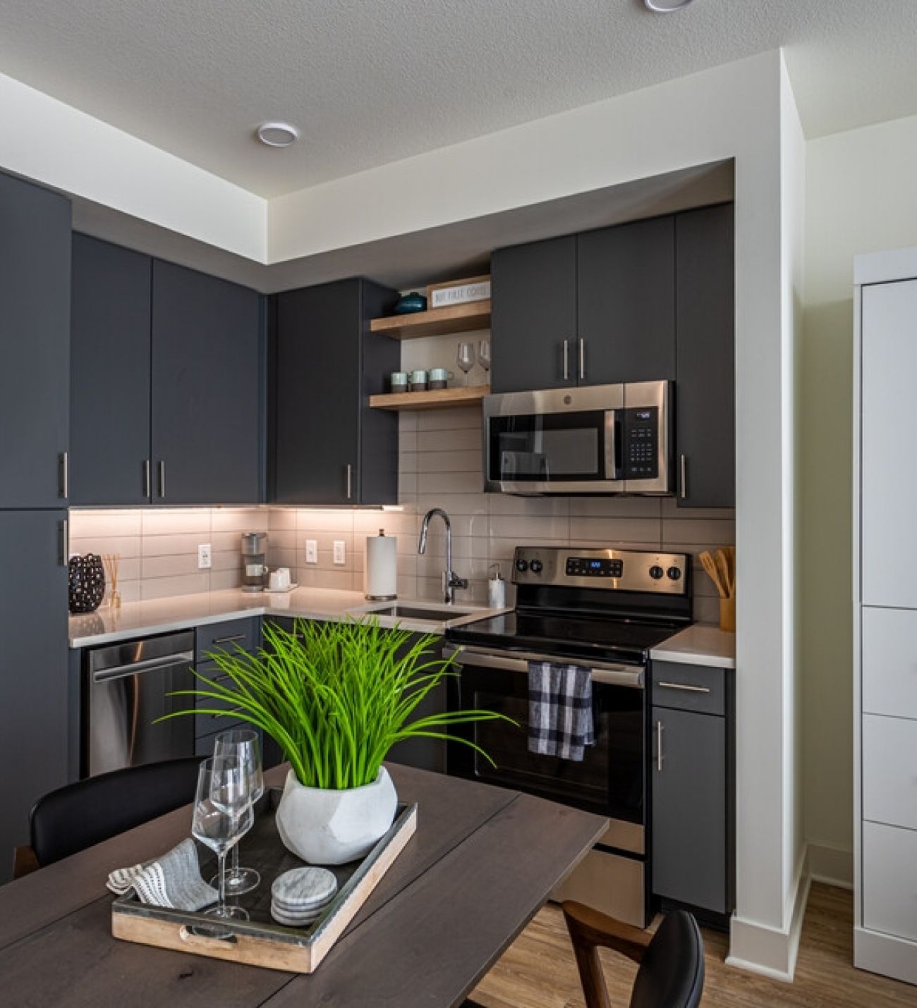 225 Sycamore Modern kitchen with dark cabinets, stainless steel appliances, and under-cabinet lighting. A small potted plant and two glasses on a tray sit on a kitchen table in the foreground. Shelves with various items are above the countertop. Bright and neatly organized space.