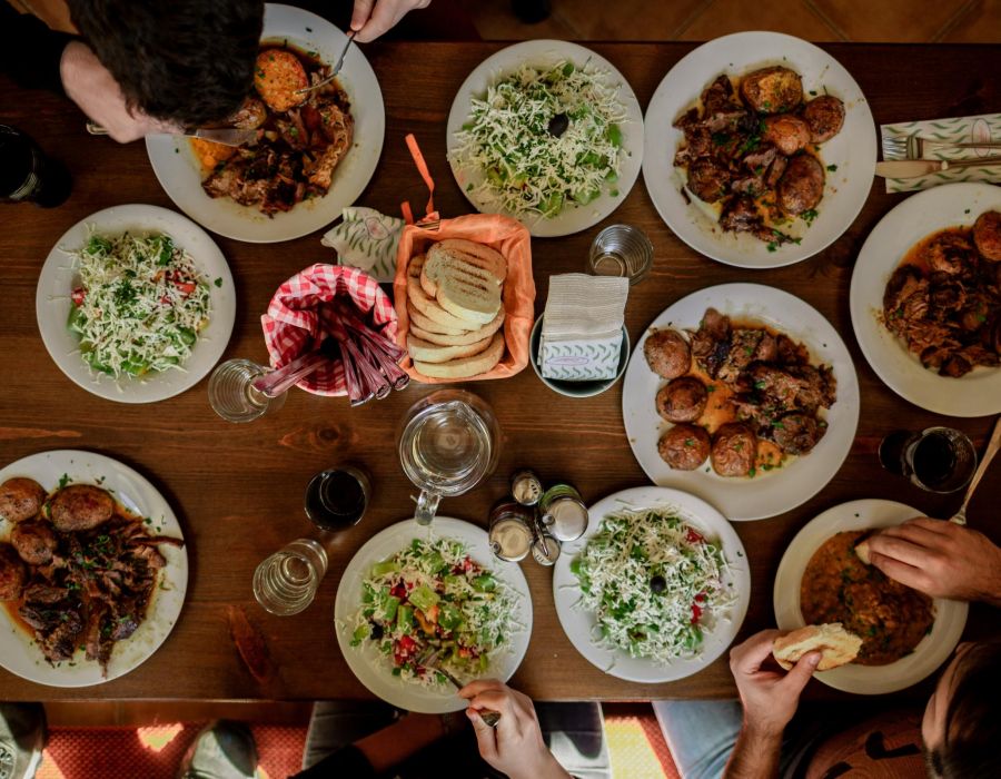 225 sycamore Top view of a dining table with several plates of food shared by a group of people. The table is filled with dishes, including salads and meat, along with glasses and a central basket of bread. The group’s hands are seen reaching for the food.