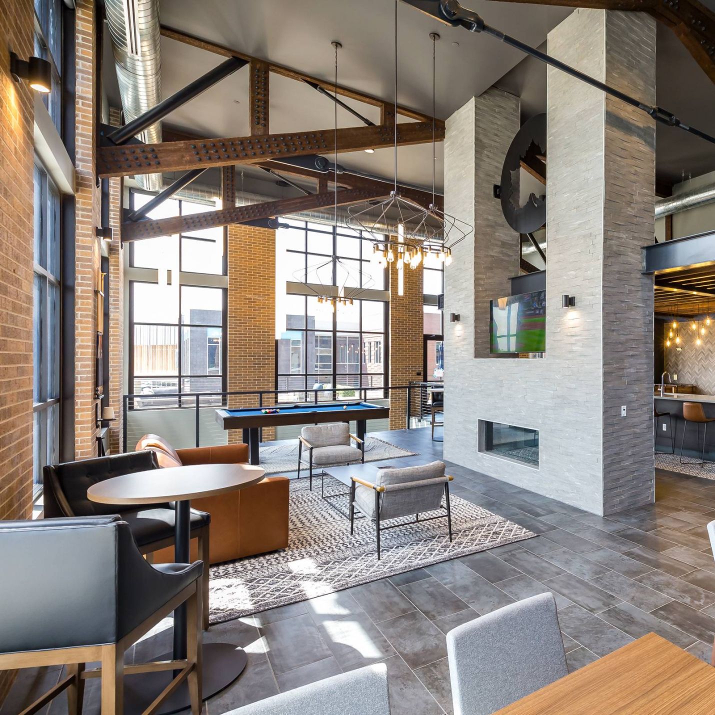 225 Sycamore modern industrial-style club room that features high ceilings with exposed beams, large windows allowing ample natural light, a central fireplace, a mix of seating areas, and a kitchen area with string lights hanging above the counter.