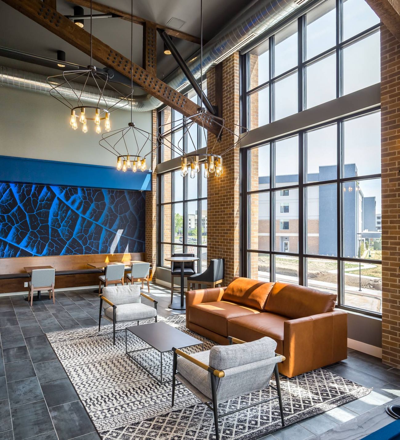 225 Sycamore lounge area features a brown leather sofa, two gray armchairs, and a wooden coffee table on a patterned rug. The room is illuminated by large floor-to-ceiling windows and stylish chandeliers, with blue accent walls and exposed beams adding architectural interest.