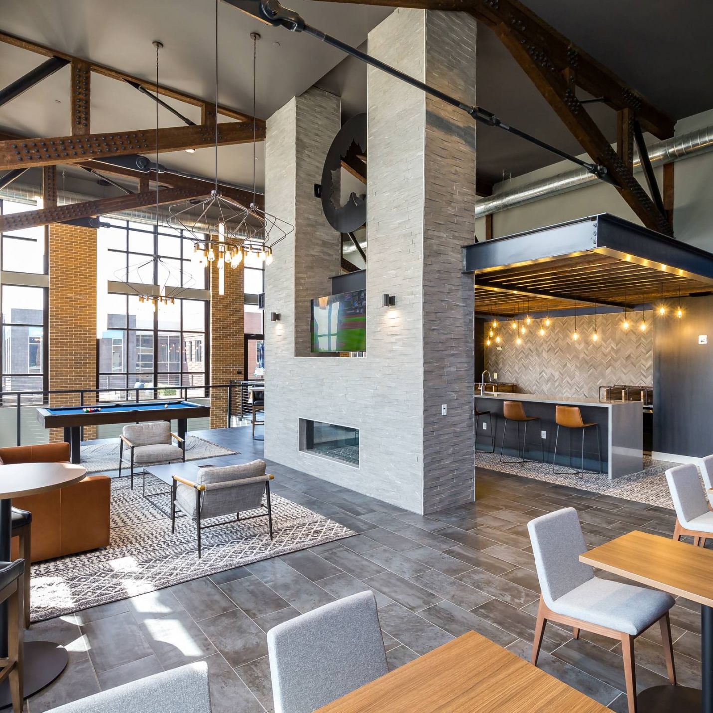 225 Sycamore modern industrial-style club room that features high ceilings with exposed beams, large windows allowing ample natural light, a central fireplace, a mix of seating areas, and a kitchen area with string lights hanging above the counter.