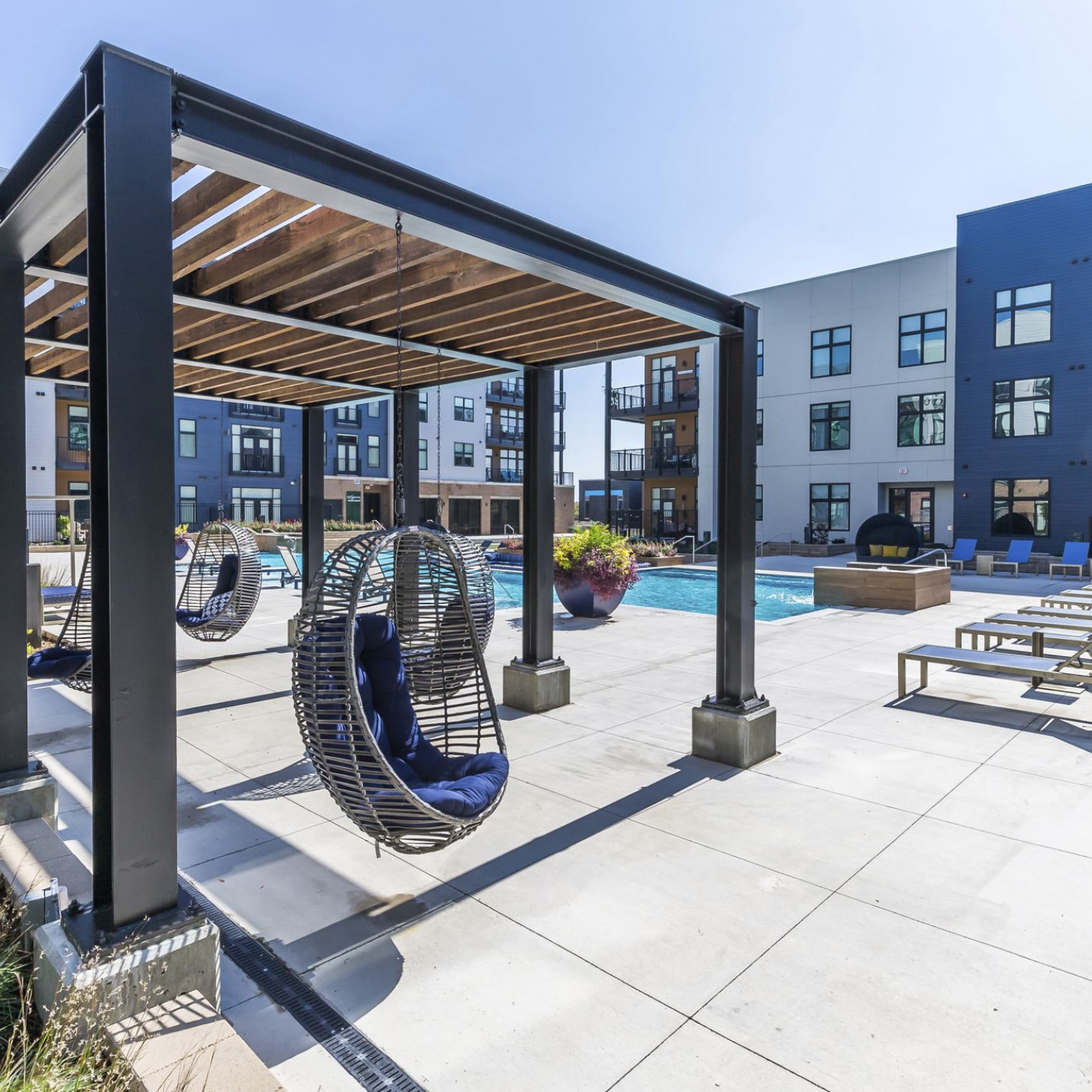 225 Sycamore Outdoor pool area with white and gray concrete flooring, a black wooden pergola, and hanging basket chairs with blue cushions. The background features modern apartment buildings with large windows, lounge chairs, and potted plants near the pool.